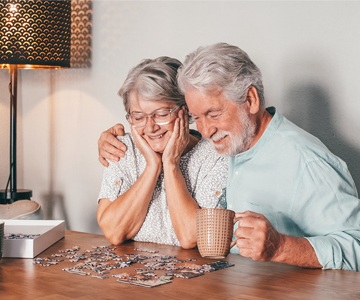 Elderly man has arm around elderly smiling woman as they sit next to each other and analyzes a puzzle.