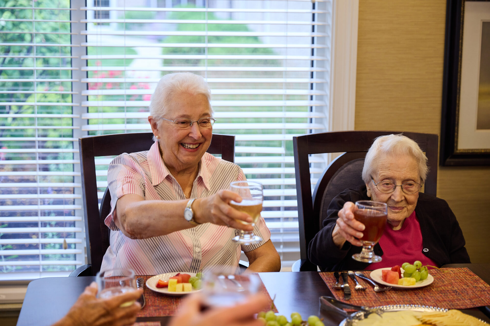 Two senior women holding their beverage glasses up for a cheers, smiling and sitting around a dinner table.