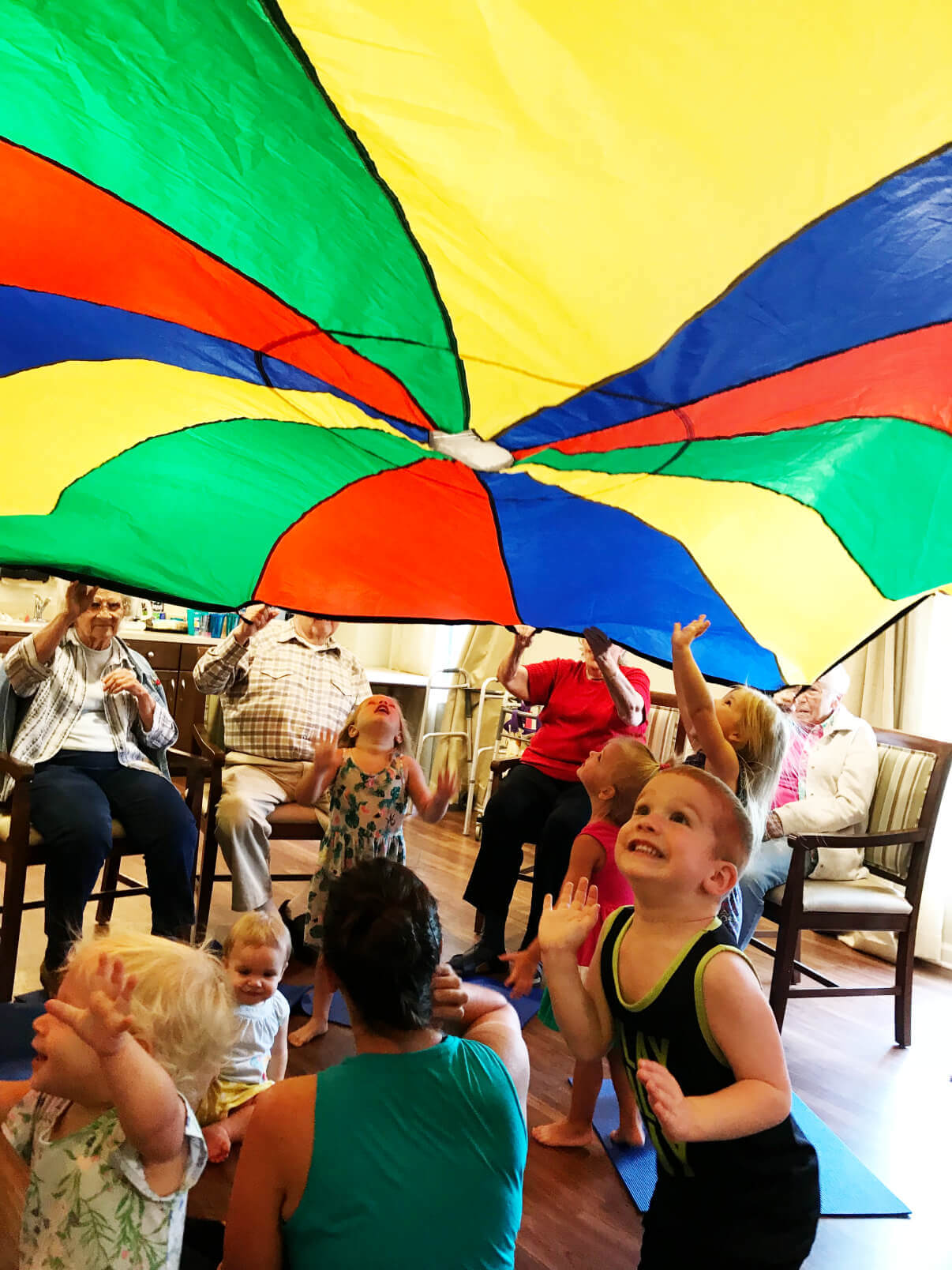 Residents and children play with a colorful parachute in a senior living community room.