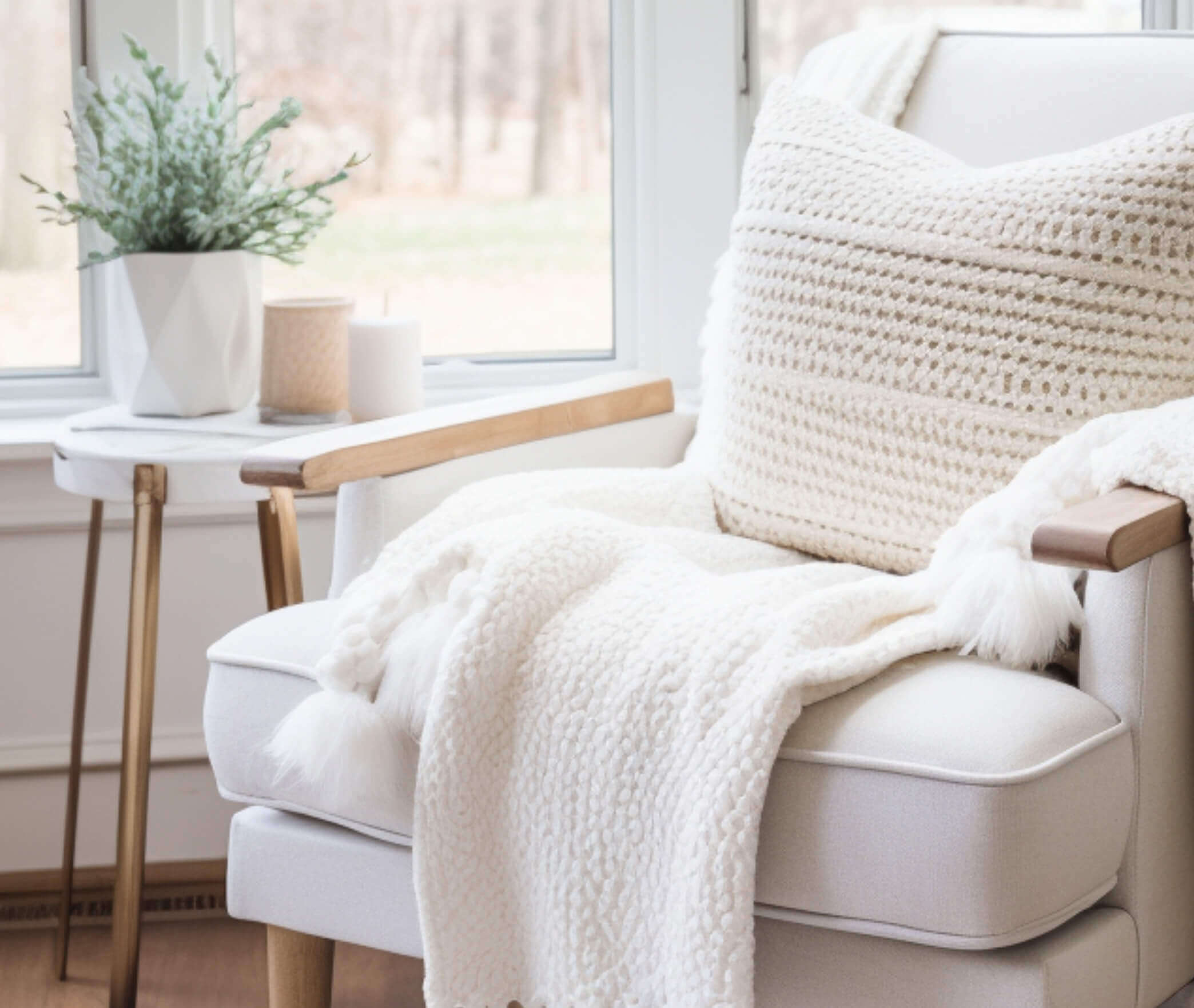 Cozy beige chair with knitted pillow and throw blanket by a window in natural light.