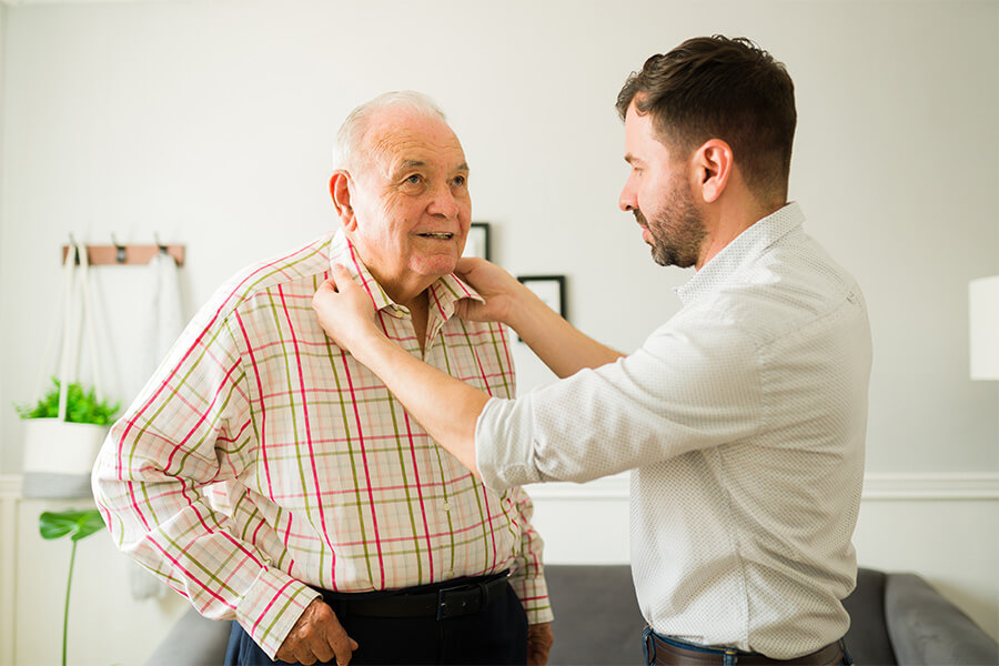 Caregiver assisting a senior man in a plaid shirt in a living space.