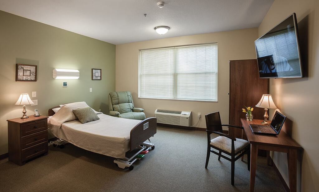 Cozy bedroom in senior community with bed, chair, desk, and TV.