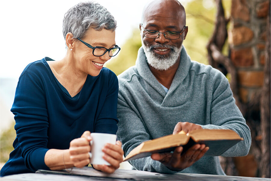 Elderly couple enjoys reading a book and coffee outdoors in a serene setting.