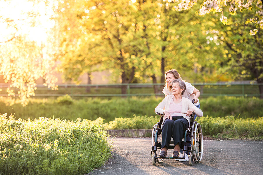 Smiling elderly woman in wheelchair being pushed by a caregiver in a sunny park.