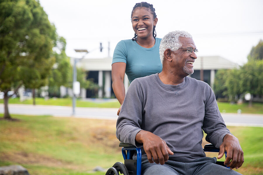 Woman smiling while pushing elderly man in wheelchair outdoors.