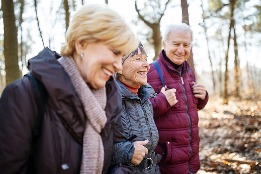 Three seniors enjoy a walk in a wooded area, smiling and dressed in warm jackets.