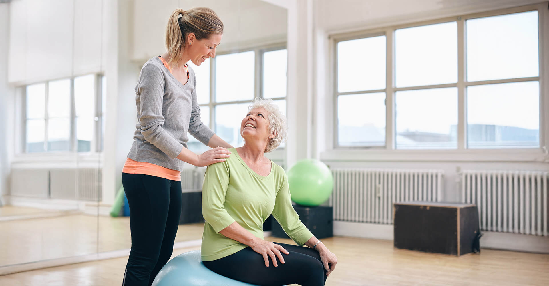 Young woman assisting senior woman on exercise ball in a sunny room.