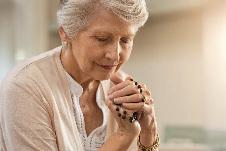 Elderly woman in peaceful prayer holding rosary with closed eyes in soft light.