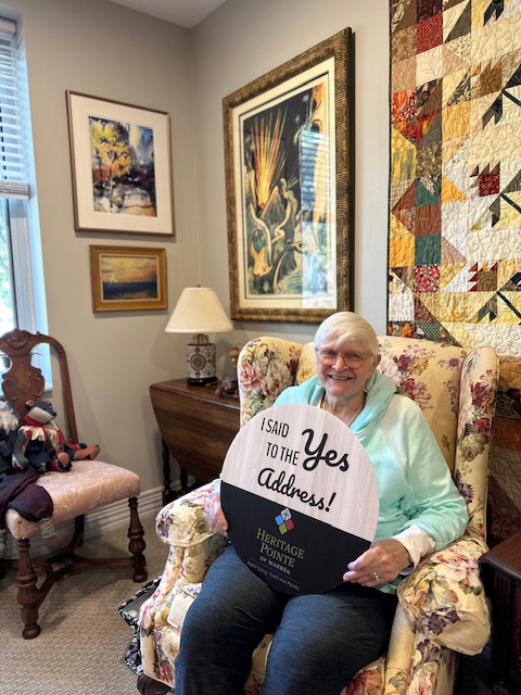 Jocelyn Smith holding a "I said yes to the address" sign in her home decorated with homemade quilts
