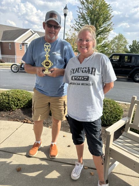 Two people holding a trophy outside a community building with parked cars and greenery.