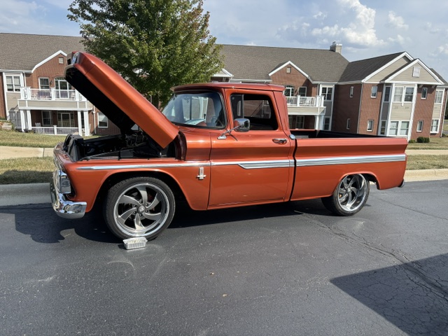 A classic red pickup truck is parked with its hood open in front of residential units on a sunny day.