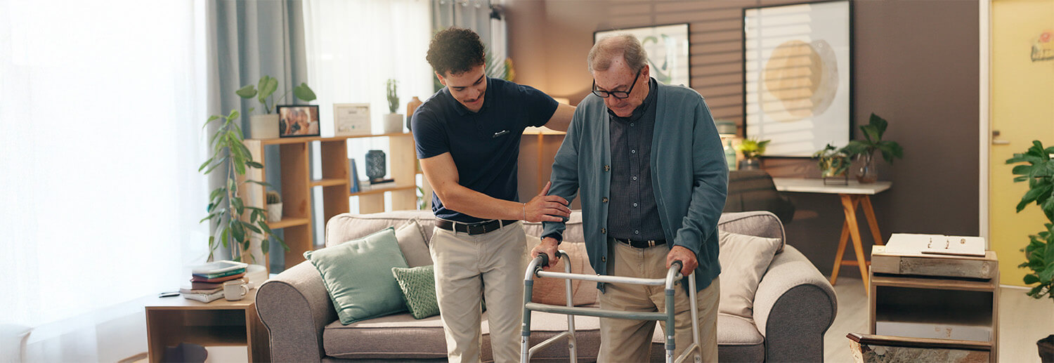 Senior man uses walker while being assisted by a younger man